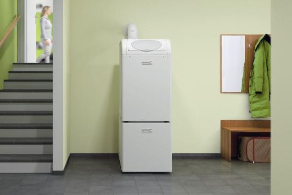 Eine weiße Heizungseinheit steht im Flur neben einer Treppe und Garderobe mit grünem Mantel; die Wände sind hellgrün gestrichen.